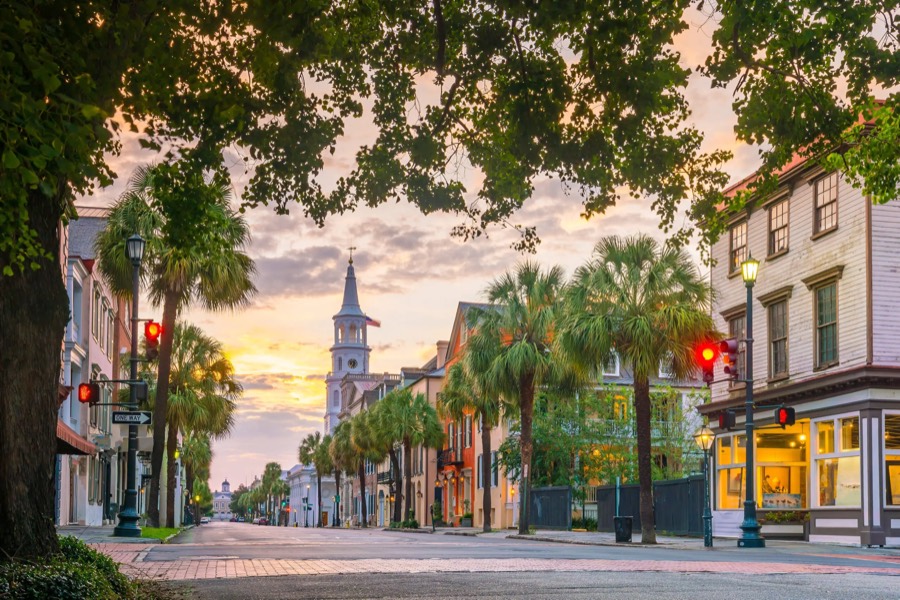 A Charleston street at dusk lined with palmetto trees and historic buildings, a white church steeple against the sunset