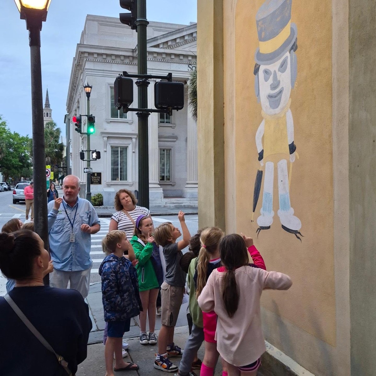 Young guests on the Junior JWalkers tour looking up and pointing at the Little Man mural painted high on a Charleston street corner, with their guide beside them