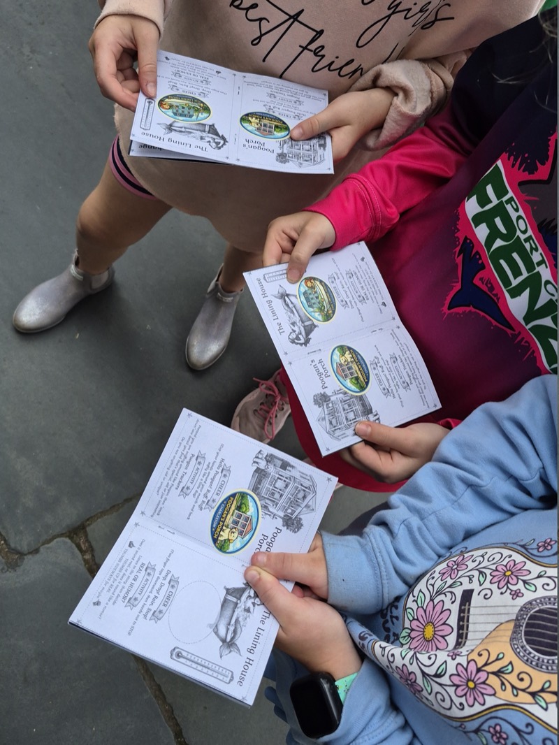 Three children on a Charleston sidewalk holding open their Junior JWalker passports, each page showing an illustrated stop — Pogue's Porch and The Lining House — with a colour sticker earned beside it