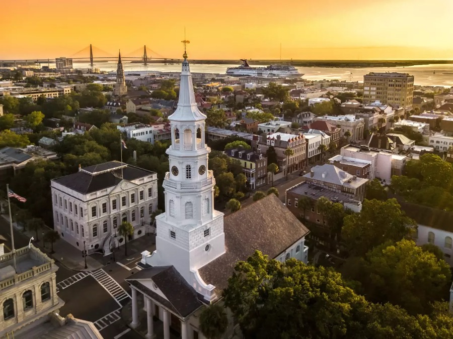 Aerial view of Charleston at sunset: a white church steeple rising above the historic district, with the harbour, the Ravenel Bridge and a cruise ship beyond